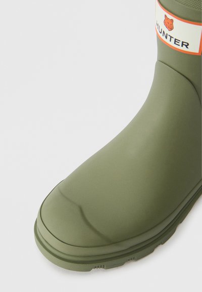Bota de borracha verde com bico arredondado, textura suave, e um rótulo branco com o nome da marca "HUNTER" e um logo de urso.