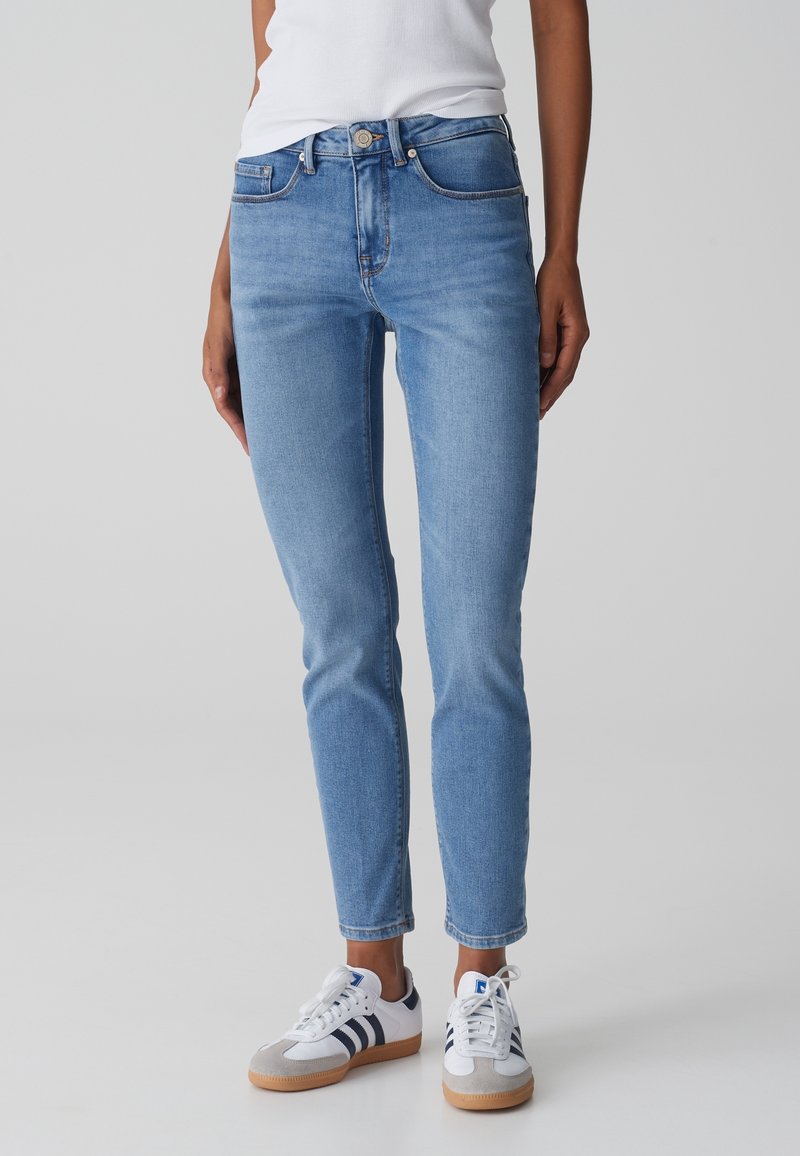 Jean slim fit bleu clair porté avec des baskets blanches à rayures noires, associé à un haut sans manches blanc, debout sur un fond uni.