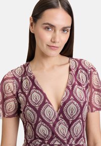 Wickeldress in Burgunderrot mit cremefarbenen Blumenmustern. Besitzt einen tiefen V-Ausschnitt und kurze, transparente Ärmel. Glatter Stoff.