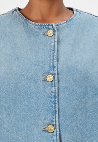 Lichtblauwe denimjacket met een ronde halslijn, vier gouden knopen en een eenvoudig ontwerp. De stof heeft een licht gestructureerde afwerking.