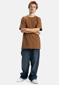 Brązowy bawełniany t-shirt o luźnym kroju, z krótkim rękawem i naszywką z logo, zestawiony z luźnymi niebieskimi dżinsami i białymi trampkami.