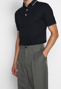 Svart texturerad polo med ett mönster av små hjärtan, vit kant på kragen och ärmarna, matchad med grå skräddarsydda byxor.