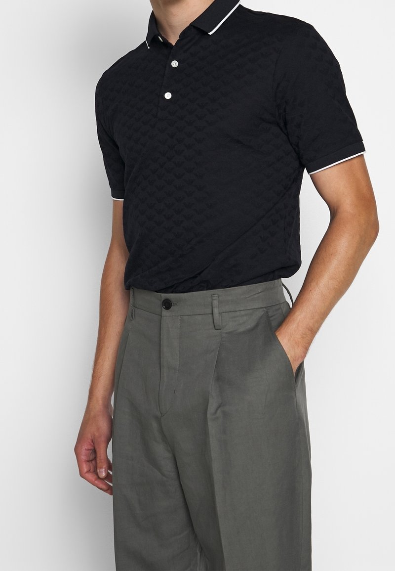 Svart texturerad polo med ett mönster av små hjärtan, vit kant på kragen och ärmarna, matchad med grå skräddarsydda byxor.