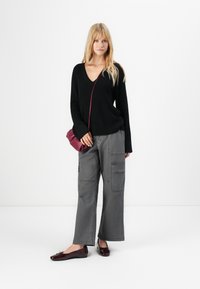 Woman in black V-neck sweater, gray wide-leg cargo pants, burgundy flats, and matching crossbody bag standing against white background.
