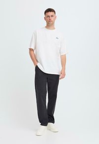 White cotton t-shirt with a light blue logo, paired with black pants and white sneakers. The fit is relaxed with short sleeves.