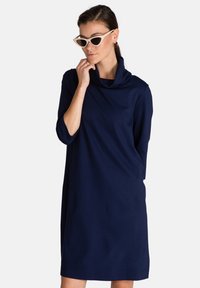 Vestido azul marino con cuello de drapeado, mangas tres cuartos y textura suave. Presenta un corte relajado y cae por encima de la rodilla.
