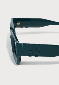 Zaļas plastmasas saulesbrilles ar biezām rāmī, ģeometrisku formu un izspiestu "HUGO" logo uz tempļiem. Spīdīga virsma ar gludu tekstūru.