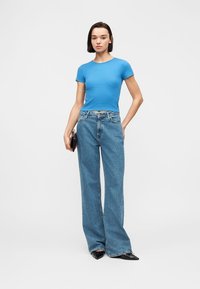 Γυναίκα που φοράει εφαρμοστό μπλε t-shirt, ψηλόμεσα φαρδιά μπλε τζιν και μαύρα μυτερά παπούτσια, στέκεται με το ένα χέρι στην τσέπη.