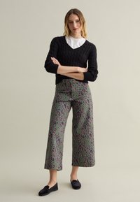 Schwarzer Kabelstrickpullover mit V-Ausschnitt, kombiniert mit weit geschnittenen grünen Hosen mit einem mehrfarbigen Leopardenmuster. Schwarze Slipper.
