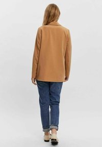 Vaaleanruskea bleiseri, jossa on käänteet, istuva leikkaus ja sileä kangas. Yhdistetty rentoihin sinisiin farkkuihin, joissa on käännetyt hihat ja vaaleat kengät.