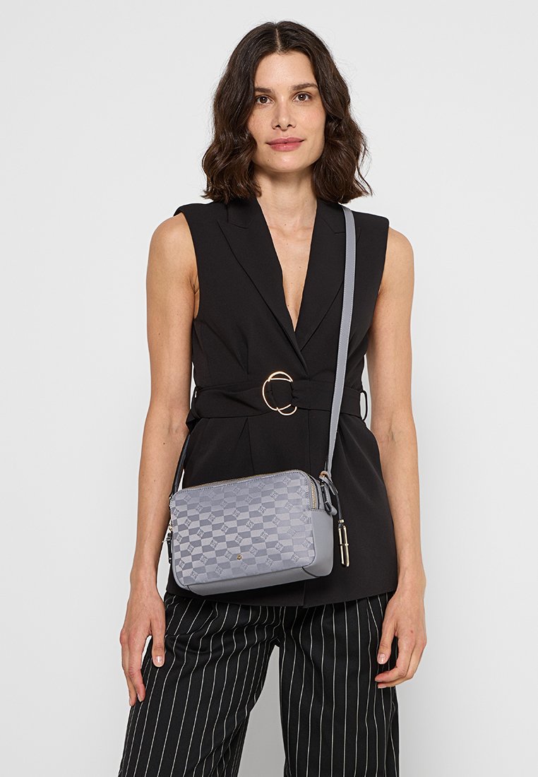 Grå mönstrad crossbody-väska med rektangulär form, texturerat material, dragkedjestängning och justerbar rem, hålls av en modell i en svart topp.