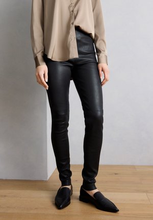 Άτομο που φοράει μπεζ μακρυμάνικη μπλούζα, μαύρο δερμάτινο κολάν και μαύρα μοκασίνια με μυτερή μύτη, στέκεται σε ξύλινο πάτωμα μπροστά σε γκρι τοίχο.