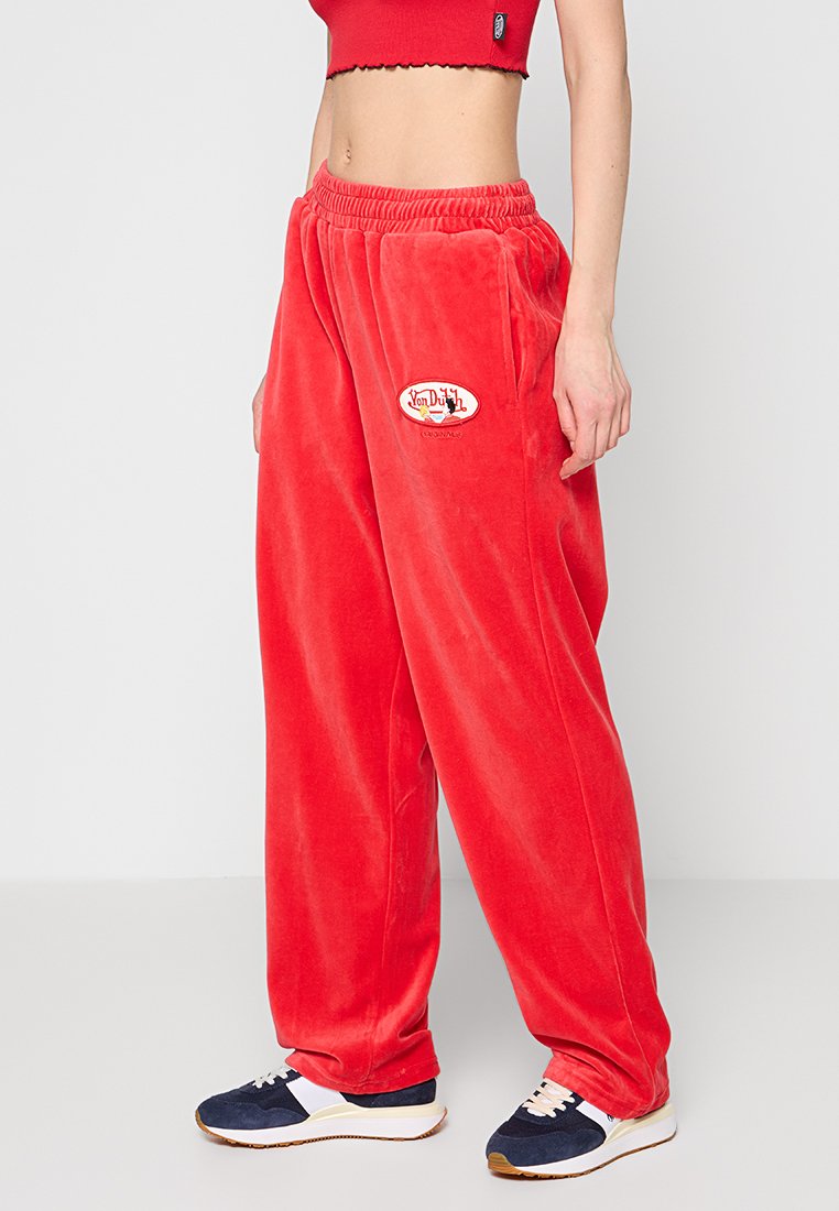 Rote Samt-Sweatpants mit elastischem Bund, ausgestattet mit einem runden Logo-Patch. Lockere Passform und weiche Textur, kombiniert mit navyblauen und weißen Sneakern.