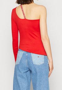 Kobieta w czerwonej bluzce na jedno ramię z paskiem Tommy Jeans oraz jasnoniebieskich, wysokich dżinsach z dwiema tylnymi kieszeniami.