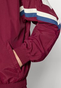 Rękaw bordowej kurtki z białymi i niebieskimi paskami, elastycznym mankietem, a dłoń częściowo wsunięta do przedniej kieszeni na neutralnym tle.