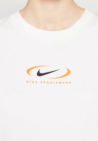 White sweatshirt featuring a black Nike logo and an orange oval accent, with ribbed collar and soft texture.