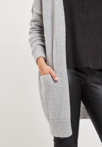 Světle šedý kardigan z měkkého, texturovaného materiálu, s kleslými ramenními švy a přední kapsou. Nositý přes černý top a kalhoty.