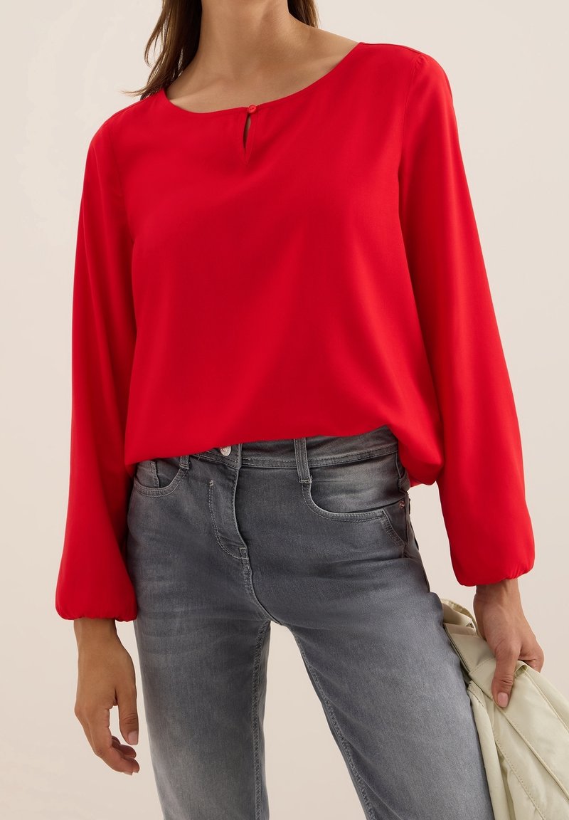 Femme portant un chemisier rouge ample à manches longues avec un petit bouton près de l'encolure, associé à un jean gris délavé, tenant une veste légère.