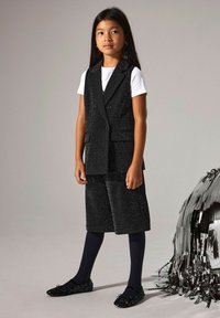 Črno, bleščeče dvodelno oblačilo z dvorednim brezrokavnikom in kratkimi hlačami do kolen, v kombinaciji z mornarsko modrimi najlonkami in okrašenimi črnimi balerinkami.