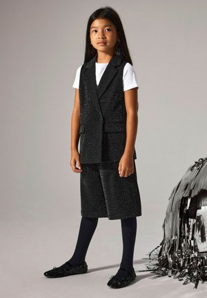 Crna, sjajna dvodijelna odjeća koja se sastoji od dvostruko kopčanog prsluka i kratkih hlača do koljena, uparena s tamnoplavim najlonkama i ukrašenim crnim balerinkama.