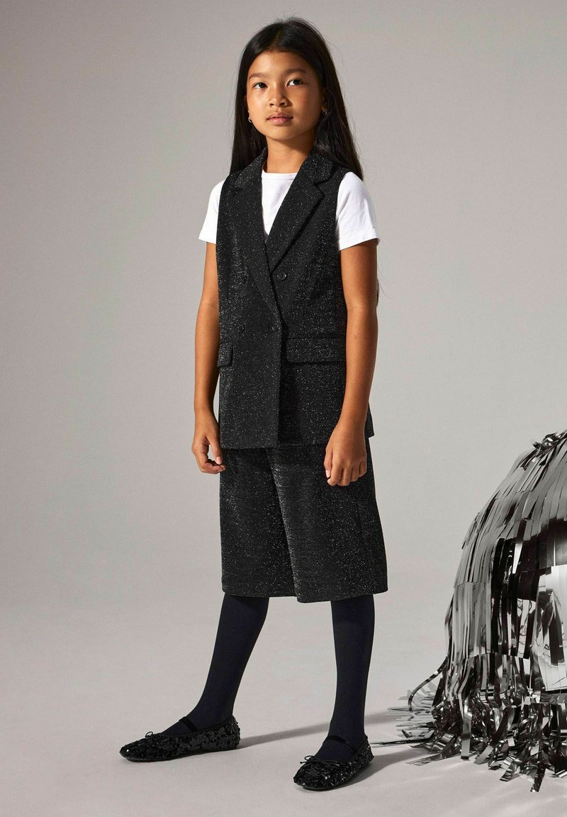 Črno, bleščeče dvodelno oblačilo z dvorednim brezrokavnikom in kratkimi hlačami do kolen, v kombinaciji z mornarsko modrimi najlonkami in okrašenimi črnimi balerinkami.