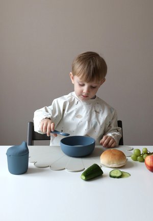 Tigela azul de plástico sobre uma mesa clara, com uma criança a mexer com uma colher azul. Por perto estão uvas verdes, pepino, uma maçã e um pão.