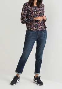 Blusa floreale a maniche lunghe con sfondo scuro, abbinata a jeans blu con risvolto e scarpe da ginnastica nere. Outfit casual e comodo.