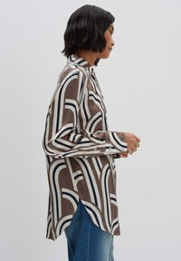 Braun- und cremefarbene Button-up-Bluse mit fließenden, geschwungenen Linien in Marineblau. Lange Ärmel mit Knopfmanschetten, Seitenschlitze und eine lockere Passform.