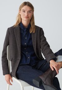 Blazer a quadri marrone sopra una camicia in denim scuro, abbinato a pantaloni in denim scuro e stivali con tacco marroni. Texture morbide e vestibilità su misura.