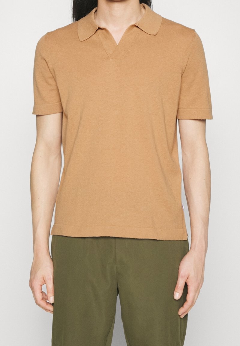 Personne portant un polo manches courtes couleur camel avec un col et un pantalon vert, debout devant un fond clair uni.