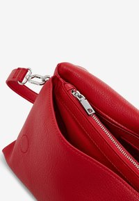 Borsa a mano in pelle rossa con superficie testurizzata, scomparto principale con zip e hardware argentato. Il design presenta una forma triangolare.