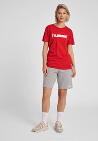 Röd bomullströja med vit "hummel"-logotyp, tillsammans med grå bomullsshorts. Skorna är beige sportskor med vita detaljer och strumpor.