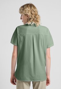 Camicia a maniche corte di colore verde chiaro, realizzata in tessuto testurizzato, con un plissettatura sul retro e una vestibilità rilassata. Design semplice senza hardware visibile.