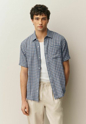 Short-sleeve shirt in navy blue with a white geometric pattern, featuring a button front and a chest pocket, worn over a white tank top.