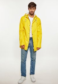 Gele waterdichte regenjas met een rits aan de voorkant, trekkoordkap, en drukknoopsluiting, gecombineerd met blauwe jeans en witte sneakers.
