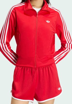 Person wearing red Adidas track jacket with white stripes and matching red shorts with white trim and Adidas logo.