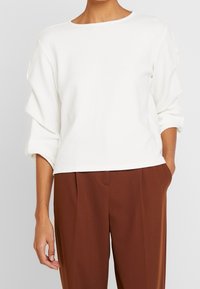 Kvinna som bär en vit långärmad texturerad topp med uppvikt ärm och bruna högmidjade plisserade byxor, med en hand i fickan.