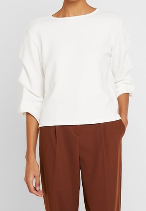 Donna che indossa una maglia bianca a maniche lunghe con texture e maniche arrotolate, abbinata a pantaloni marroni a vita alta con pieghe, con una mano in tasca.