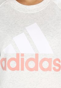 Primo piano di una felpa grigia chiara con un grande logo bianco a tre strisce e la scritta "adidas" in rosa sul petto.