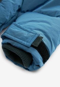 Blå pufferjakkeærme med et glat ydre materiale, tekstureret sort strikforing og en Velcro-strop til lukning.