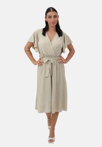 Vestido envolvente beige con mangas cortas tipo volantes, cintura elástica y detalle de lazo. Tejido suave y ligero, de longitud a la rodilla y con una silueta fluida.