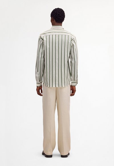 Homme de dos portant une chemise blanche à manches longues avec des rayures verticales vertes et un pantalon large beige, debout sur un fond uni.