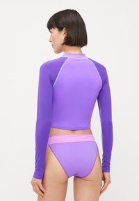Lilla langermet badetopp med kontrasterende sømmer, kombinert med matchende bikini-bunn. Stoffet har en glatt tekstur og en kroppsnær design.
