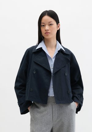 Frau mit langen schwarzen Haaren, die eine marineblaue, kurz geschnittene zweireihige Jacke, ein hellblau gestreiftes Hemd und graue Hosen trägt, die Hände in den Taschen.