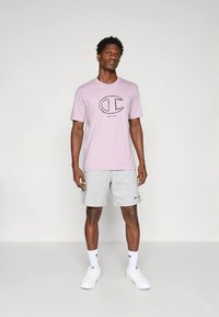 Lichtpaarse T-shirt met een zwart logo, gecombineerd met grijze korte broek. Model draagt witte sokken en schoenen, staand op een effen achtergrond.
