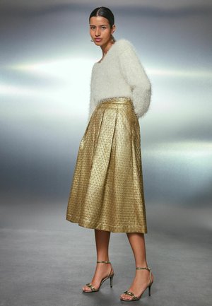 Flauschiger weißer Pullover mit langen Ärmeln, kombiniert mit einem plissierten goldenen, knielangen Rock, der ein strukturiertes Muster aufweist, und hohen Sandalen.