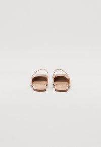 Sandales couleur nude avec une semelle plate, un dos ouvert et des sangles croisées. Fabriquées en matériau lisse avec une semelle intérieure texturée pour le confort.