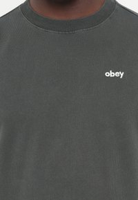 Sötét szürke pamut póló bordázott kerek nyakkal és egy kis fehér "obey" logóval a bal mellen. Puha tapintású, laza stílus.