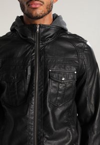 Svart läderjacka med dragkedja framtill, grå huva som är fodrad, två bröstfickor med tryckknappar och ribbade detaljer på axlarna.