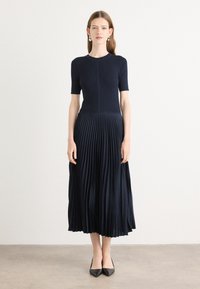 Knit navy dress with short sleeves, featuring a pleated skirt. Smooth top transitions to textured, flowing fabric. Pointed black heels.